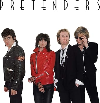 The Pretenders band members posing together in studio, featuring Chrissie Hynde in a red jacket, associated with the song Brass in Pocket.