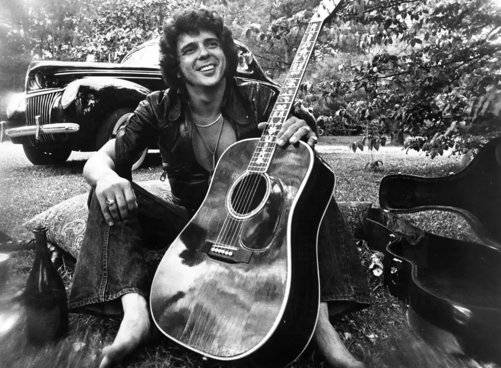 Bobby Whitlock in 1975 – Remembering the Dominos’ Co-Founder - Classic Rock Artists Bobby Whitlock Dead – barefoot and smiling with acoustic guitar in 1975, sitting on grass beside vintage car.