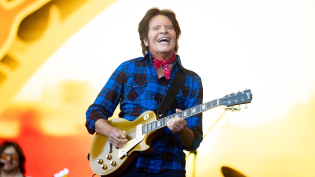 John Fogerty: 2025 BMI Troubadour Award Honoree - Classic Rock Artists John Fogerty smiling while playing guitar onstage in a portrait-style shot.