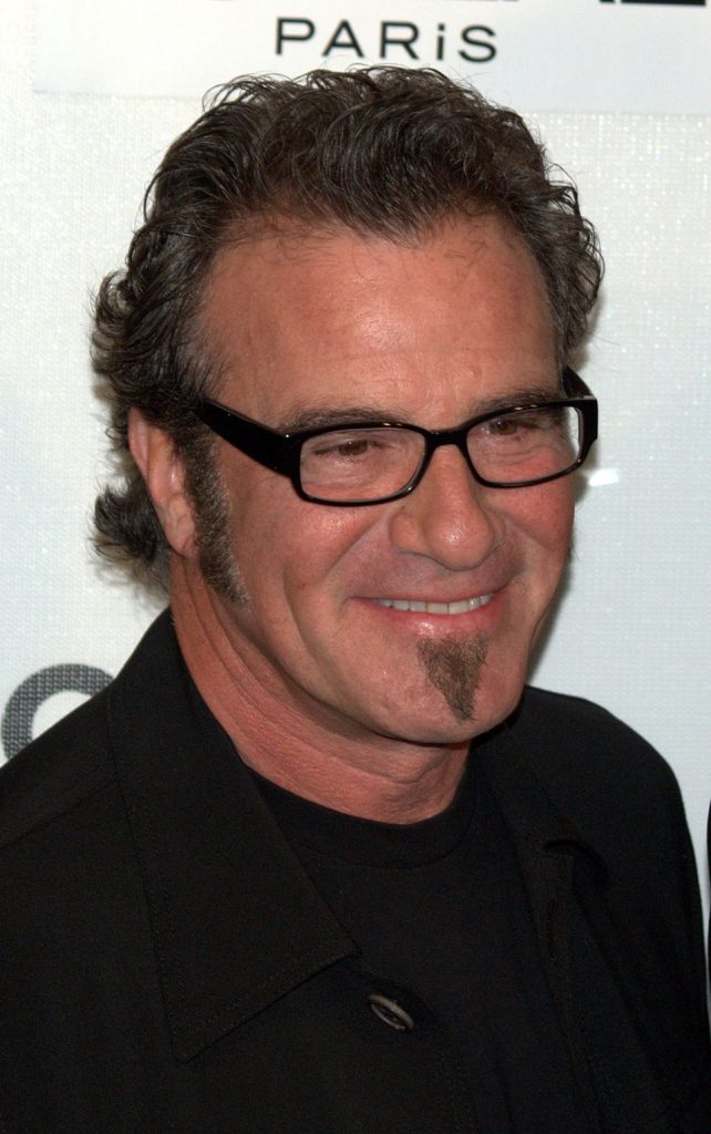 Tico Torres at Public Event - Classic Rock Artists Alt Text: "Tico Torres, drummer of Bon Jovi, smiling at a media event wearing black-rimmed glasses and a black shirt.