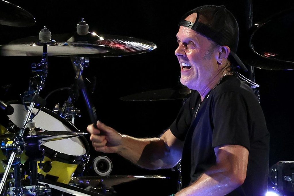  Lars Ulrich of Metallica smiling in black outfit while playing drums on stage.