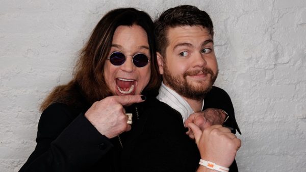 Ozzy Osbourne playfully pointing at his son Jack Osbourne, both smiling in front of a white brick wall.