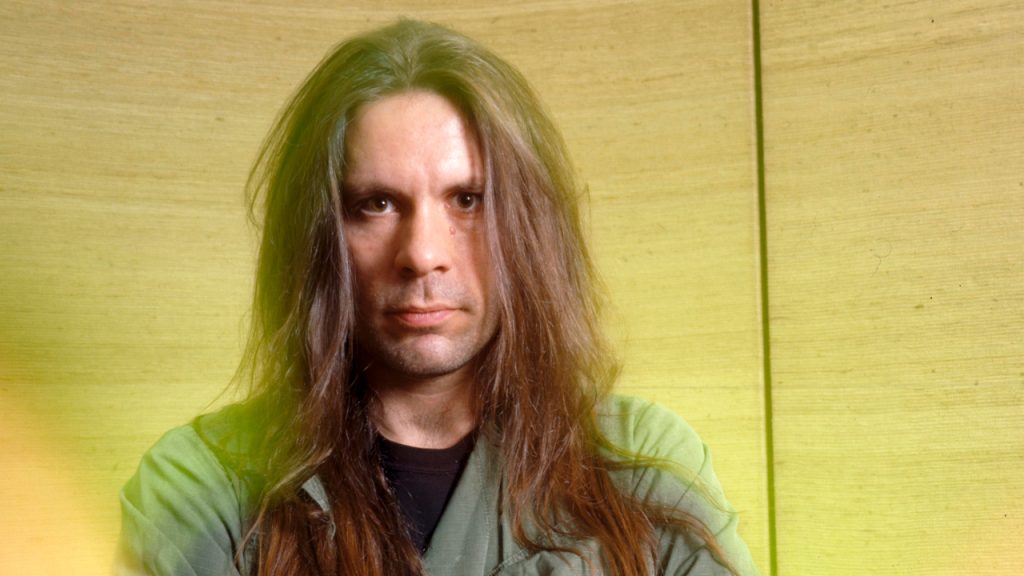 Bruce Dickinson Reflects During a Pivotal Creative Period - Classic Rock Artists Bruce Dickinson of Iron Maiden with long hair and serious expression, standing against a wooden wall background.