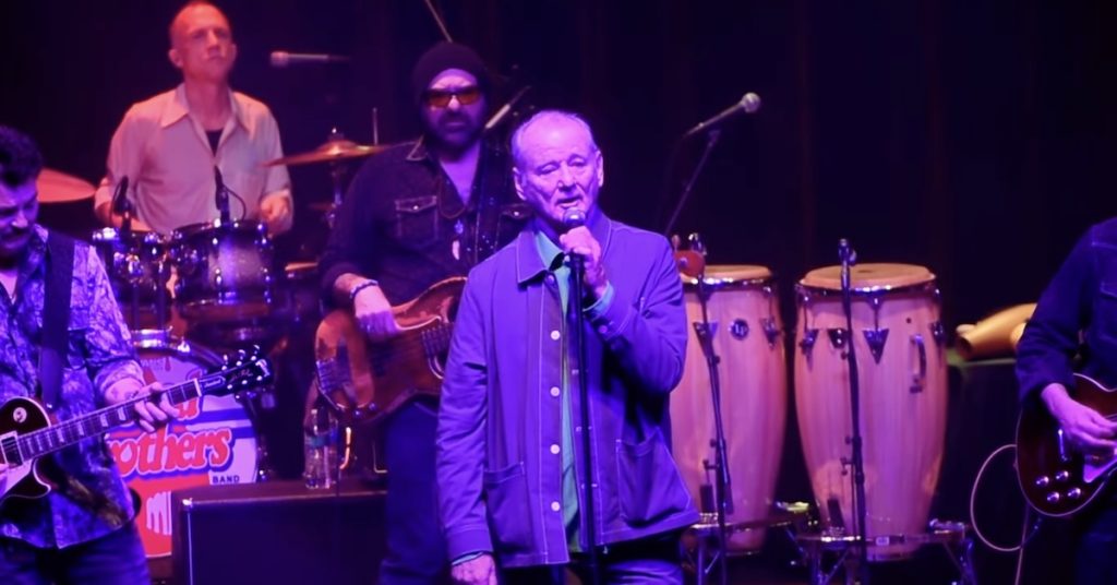 Bill Murray performing on stage with microphone in blue shirt during Bill Murray tour concert, with Blood Brothers band members playing drums and guitar in background under purple stage lighting.