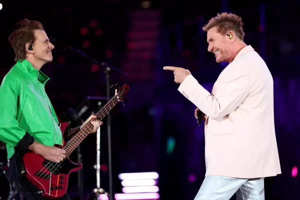 Duran Duran Band Members Perform Live on Stage During Concert - Classic Rock Artists Duran Duran Tour performers John Taylor in green jacket with red bass guitar and Simon Le Bon in white shirt pointing and smiling on stage with colorful stage lighting.