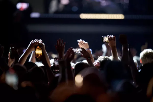 Concert crowd with raised hands and smartphones recording during a fall 2025 tour performance in a dimly lit venue.