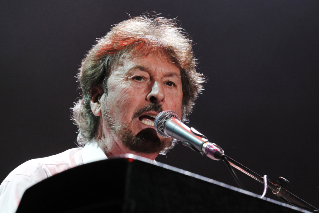Rick Davies of Supertramp performing live at keyboard with microphone in Paris, 2010, showing the legendary musician with gray hair and mustache singing into a silver microphone while playing piano