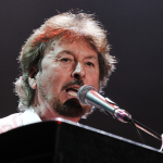 Rick Davies of Supertramp performing live at keyboard with microphone in Paris, 2010, showing the legendary musician with gray hair and mustache singing into a silver microphone while playing piano