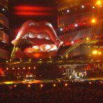 Members of the Rolling Stones performing live on stage beneath giant iconic tongue and lips logo during concert tour.