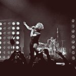 Alice in Chains performing live on stage in black and white, showing band members with bass guitar on left, lead singer at center microphone, drummer behind kit, and guitarist on right, with circular stage lighting in background.