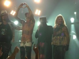 Members of Mötley Crüe performing on stage, with the shirtless drummer making a heart gesture with his hands above his head while standing between three other band members under bright stage lighting
