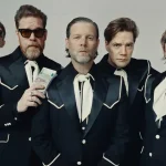 The Hives Promotional Band Portrait in Matching Suits - Classic Rock Artists A promotional photo for The Hives new album, showing the five band members standing in a line wearing matching black western-style suits and large white cravats against a grey background.