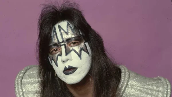 Ace Frehley in iconic Spaceman makeup featuring silver star design over his eyes and black lightning bolts on white face paint, wearing a silver sequined costume against a purple background.