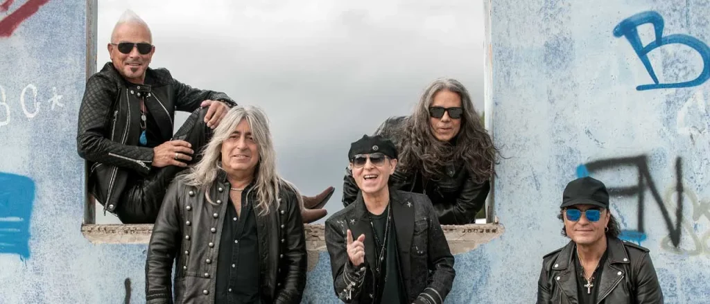 Scorpions From The First Sting 60th anniversary band photo featuring Klaus Meine, Rudolf Schenker, Matthias Jabs, Pawel Maciwoda and Mikkey Dee against graffiti wall.