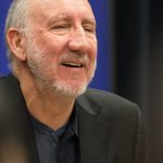 The Who Pete Townshend smiling during a public appearance, wearing a black blazer over a dark shirt.