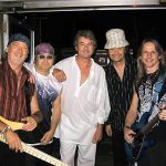 Members of Deep Purple posing together backstage in 2004, from left to right: Roger Glover holding a bass guitar, Steve Morse with sunglasses, Ian Gillan in white clothing, Don Airey wearing a striped suit and hat, and Ian Paice holding drumsticks
