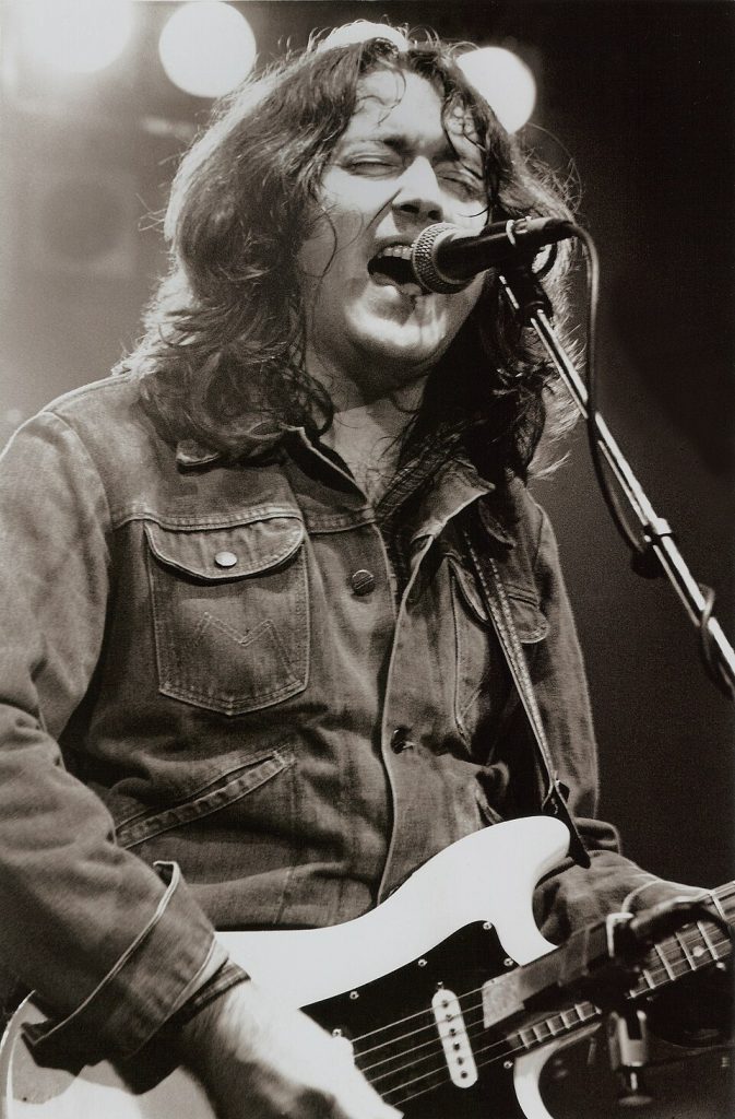 Black-and-white photo of Roy Gallagher, a long-haired musician passionately singing and playing electric guitar on stage, used for a Rory Gallagher–related article.