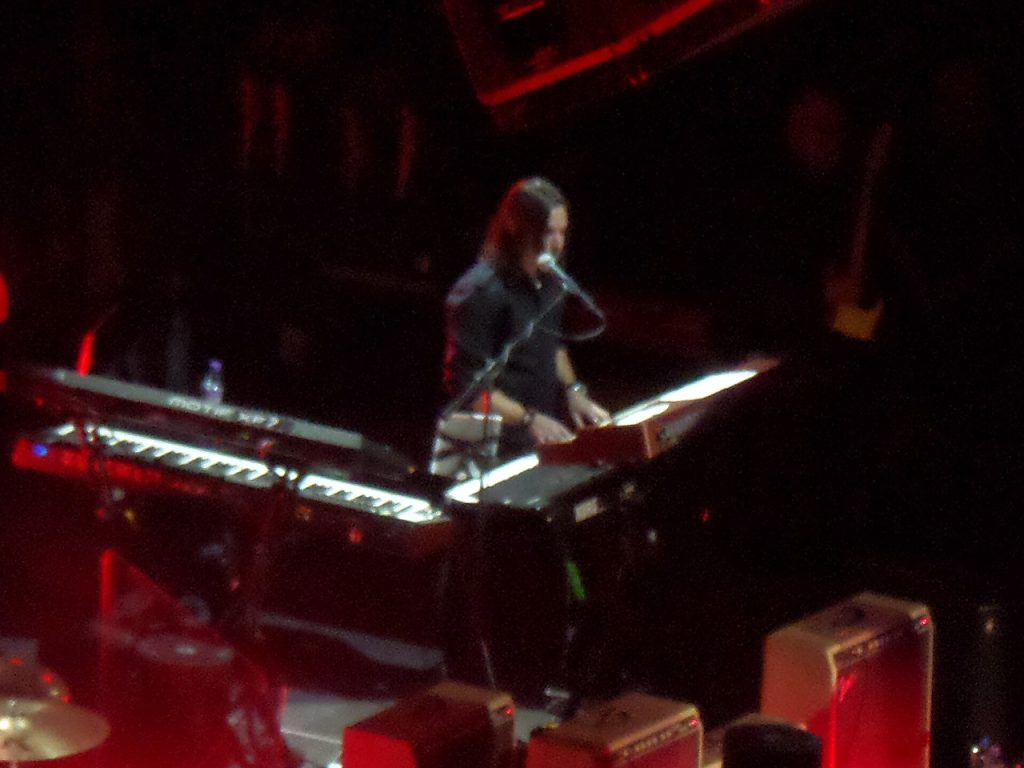 Loren Gold performing live on keyboards and vocals with The Who during a concert, surrounded by red stage lighting.