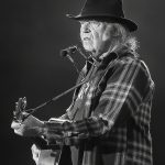 Neil Young performing live on stage in black and white, wearing a wide-brimmed hat and plaid shirt while playing acoustic guitar with harmonica rack