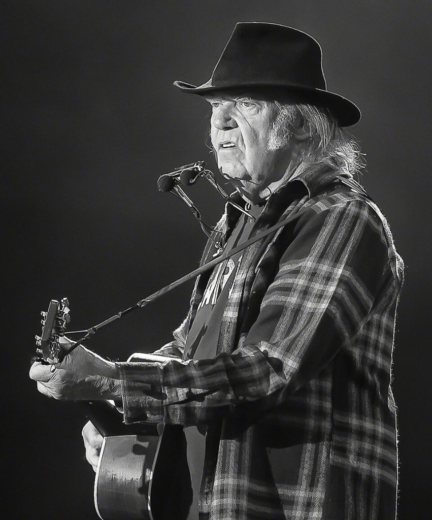 Neil Young - Biography, Albums & Latest News 2026 2 Neil Young performing live on stage in black and white, wearing a wide-brimmed hat and plaid shirt while playing acoustic guitar with harmonica rack