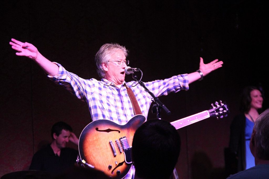 Richie Furay: The Unsung Pioneer Who Launched Country Rock and Changed Music Forever 1 Richie Furay performing live in concert in 2015, arms outstretched wide while singing into a microphone with a sunburst hollow-body guitar strapped on, wearing a blue and white checkered shirt under purple stage lighting.