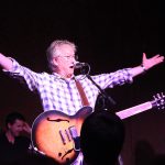 Richie Furay performing live in concert in 2015, arms outstretched wide while singing into a microphone with a sunburst hollow-body guitar strapped on, wearing a blue and white checkered shirt under purple stage lighting.