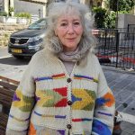 A close-up portrait of musician Suki Lahav smiling, wearing a colorful knitted cardigan with geometric patterns, on a stone street in Jerusalem with a parked car and building in the background.