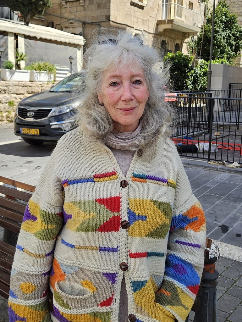 A close-up portrait of musician Suki Lahav smiling, wearing a colorful knitted cardigan with geometric patterns, on a stone street in Jerusalem with a parked car and building in the background.