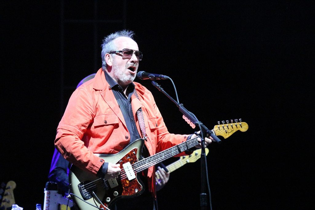 Elvis Costello performing live with his signature horn-rimmed glasses and guitar