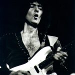 Ritchie Blackmore playing a white electric guitar during a live rock performance, captured in a dramatic black-and-white concert photograph.