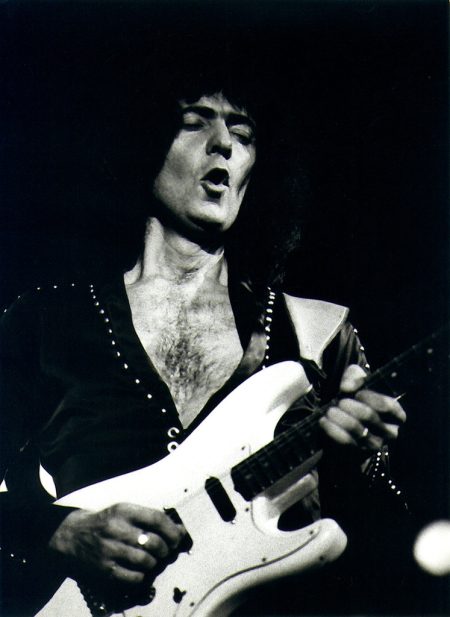 Ritchie Blackmore playing a white electric guitar during a live rock performance, captured in a dramatic black-and-white concert photograph.
